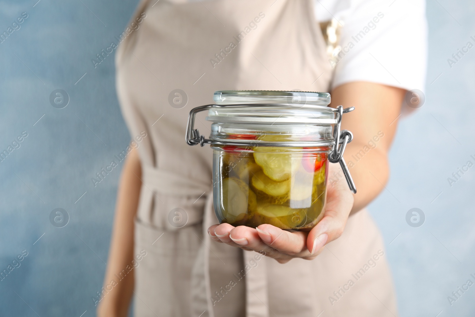 Photo of Woman holding jar of pickled cucumbers on blue background, closeup view