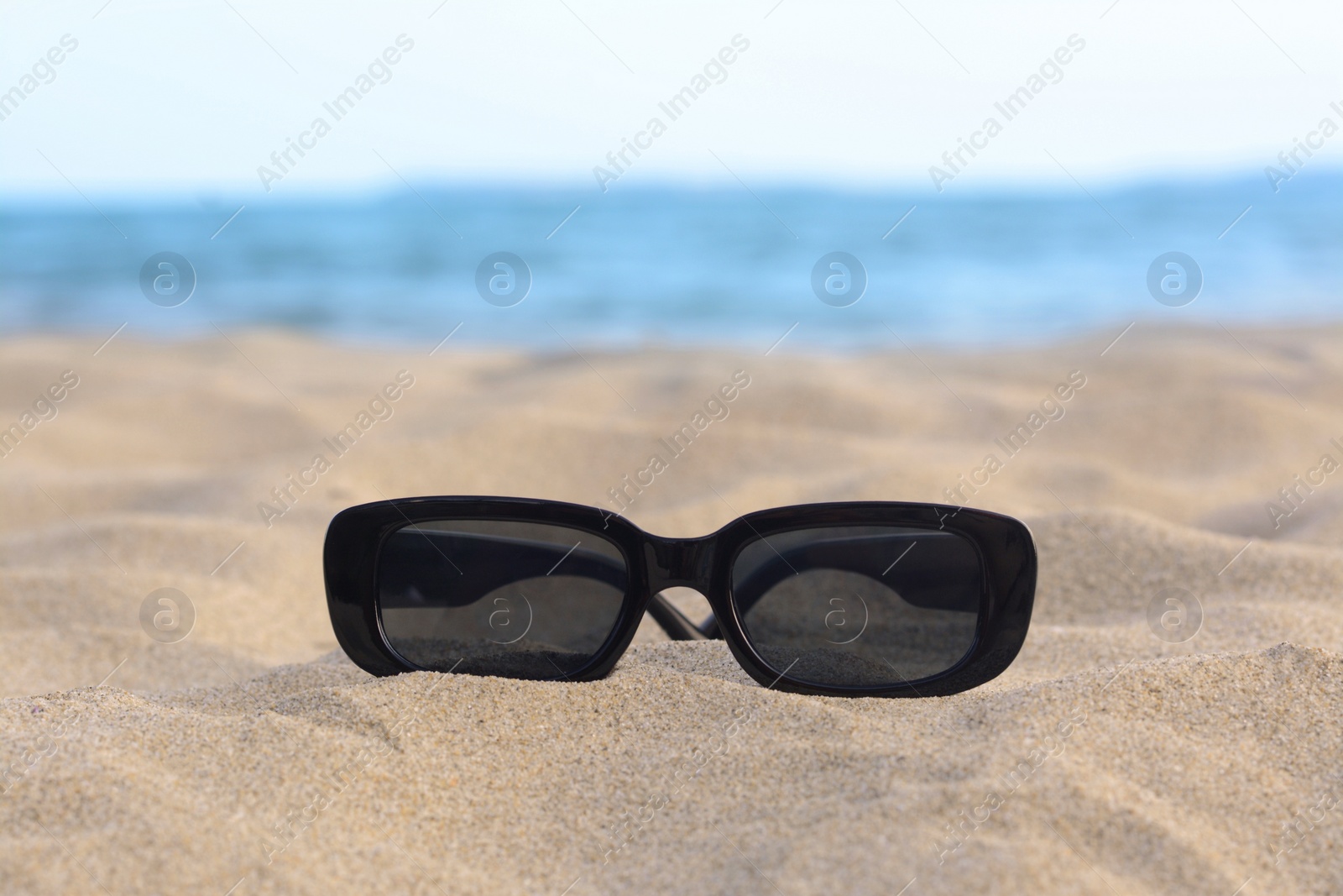 Photo of Stylish sunglasses on sandy beach near sea, closeup. Space for text