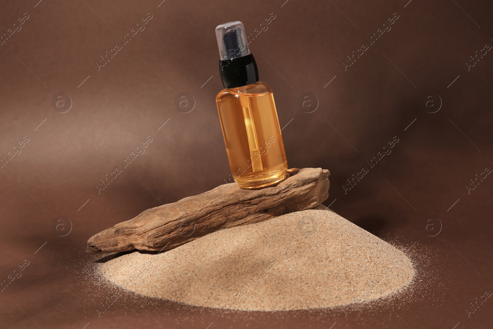 Photo of Bottle of serum and tree bark on sand against brown background