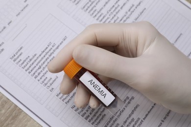 Photo of Doctor holding test tube with blood sample and label Anemia over medical form, above view
