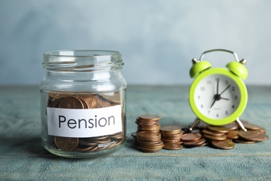 Glass jar with label PENSION and coins near alarm clock on wooden table