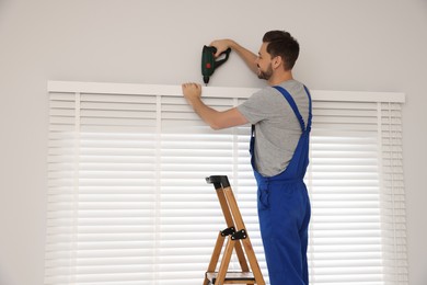 Photo of Worker in uniform installing window blind on stepladder indoors