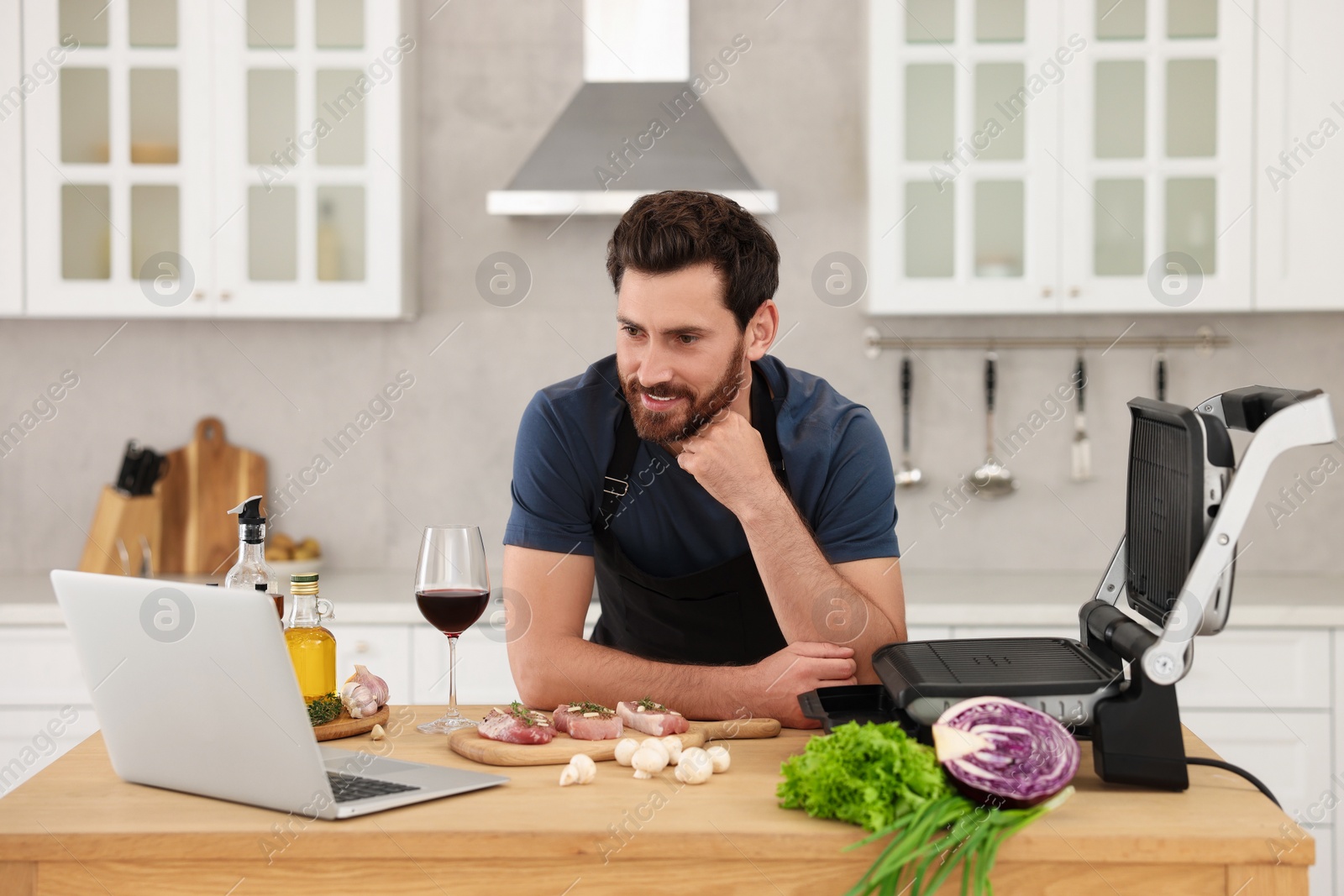 Photo of Man making dinner while watching online cooking course via laptop in kitchen