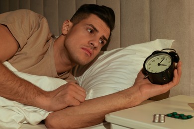 Photo of Man suffering from insomnia looking at time on alarm clock in bed indoors