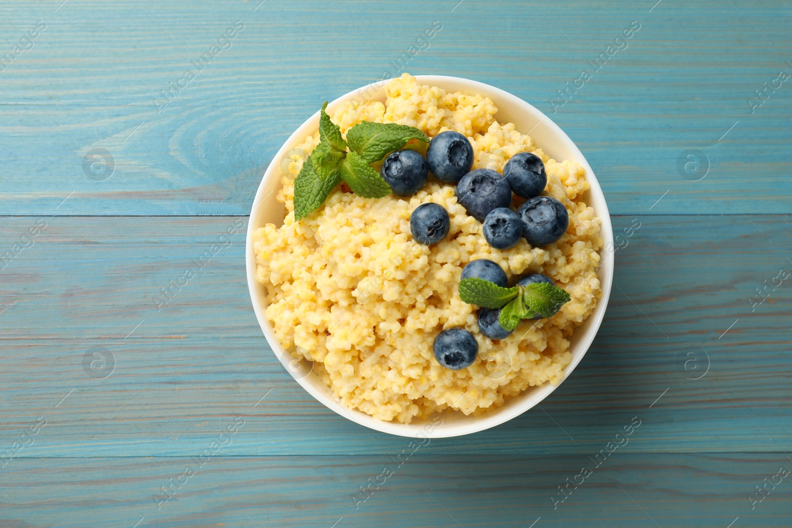 Photo of Tasty millet porridge with blueberries and mint in bowl on light blue wooden table, top view