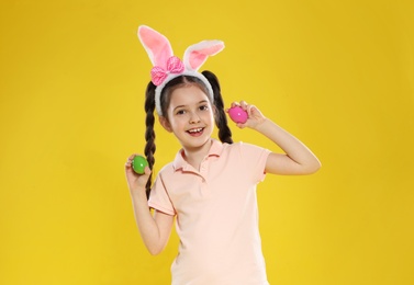 Little girl in bunny ears headband holding Easter eggs on color background