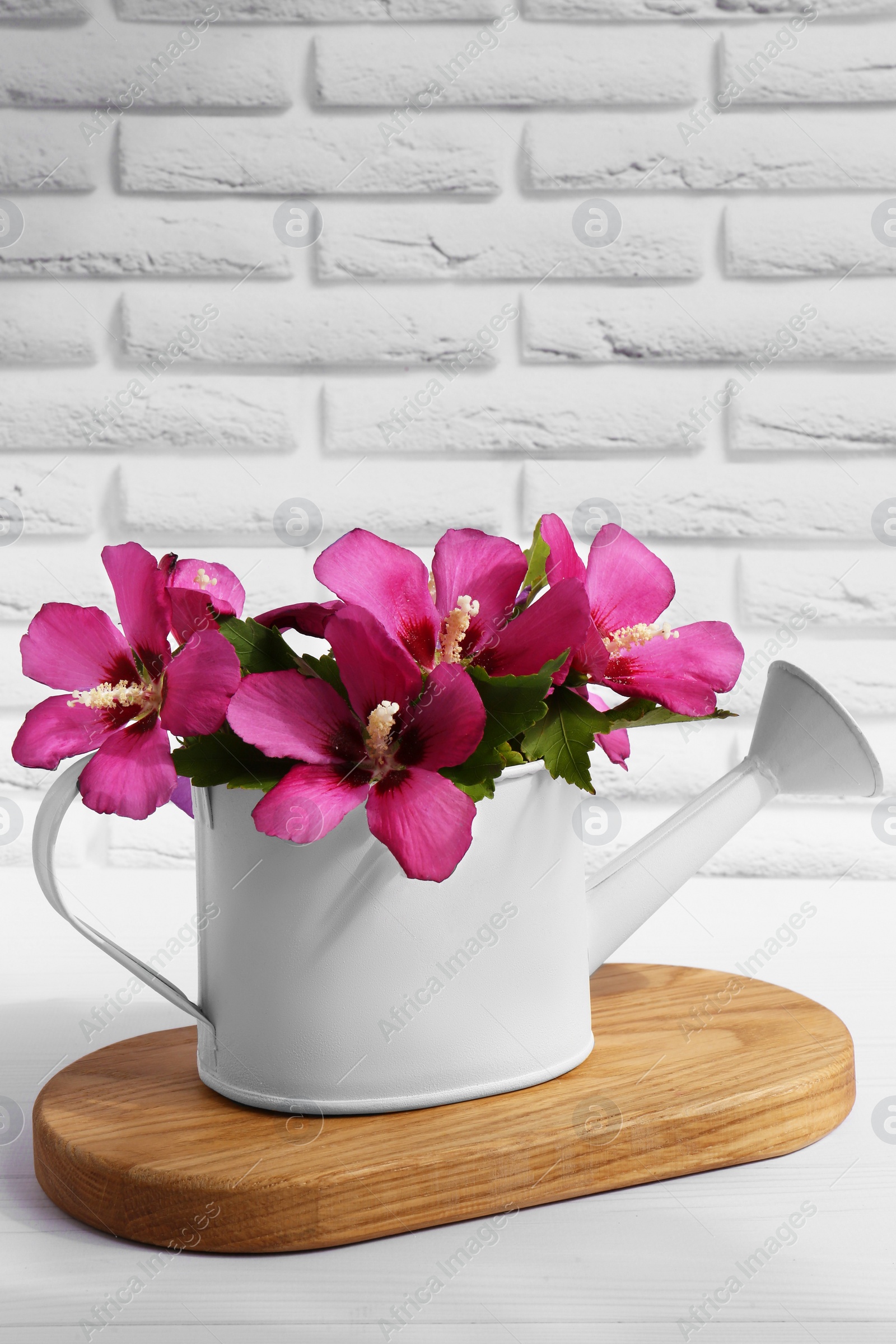 Photo of Beautiful flowers in watering can on white wooden table near brick wall