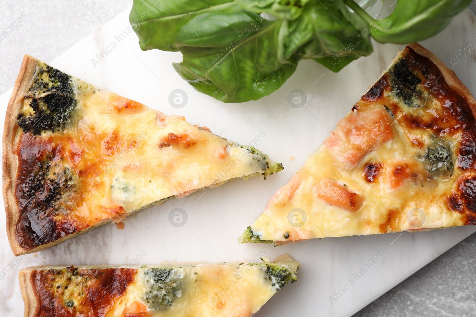 Photo of Pieces of delicious homemade salmon quiche with broccoli and basil on white board, top view