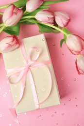 Photo of Beautiful gift box with bow, tulips and confetti on pink background, flat lay