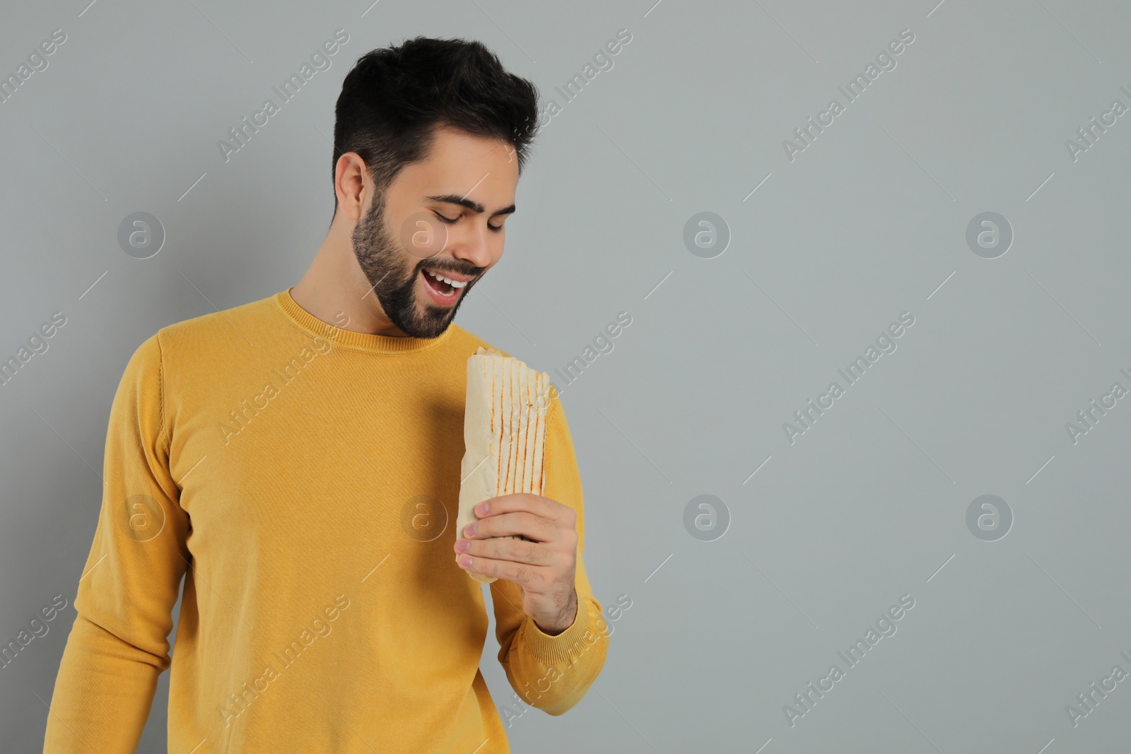 Photo of Young man eating tasty shawarma on grey  background. Space for text