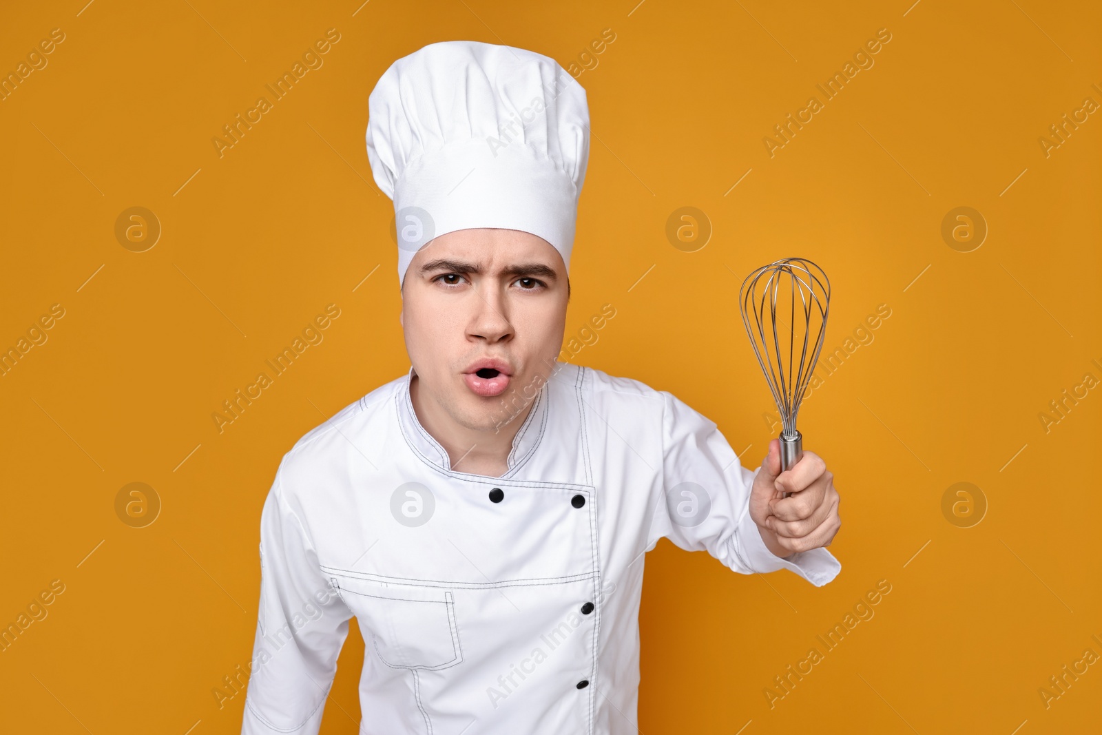 Photo of Portrait of angry confectioner in uniform holding whisk on orange background