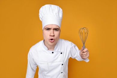 Photo of Portrait of angry confectioner in uniform holding whisk on orange background