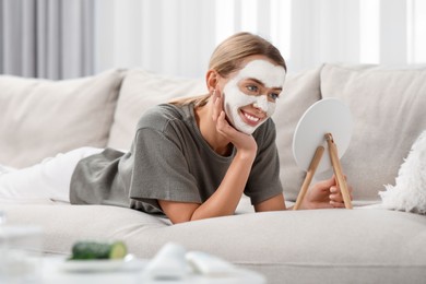 Photo of Young woman with face mask looking into mirror on sofa at home. Spa treatments