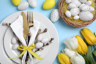 Festive table setting with tulips and painted eggs on light blue background, flat lay. Easter celebration