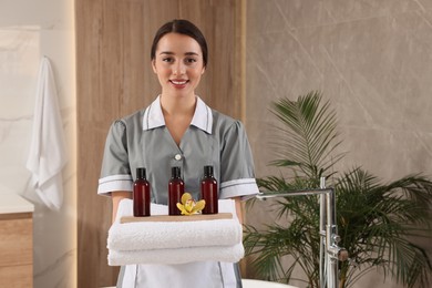 Photo of Chambermaid holding fresh towels with flower and shampoo bottles in hotel bathroom