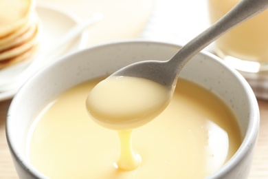 Photo of Spoon of pouring condensed milk over bowl on table, closeup. Dairy products