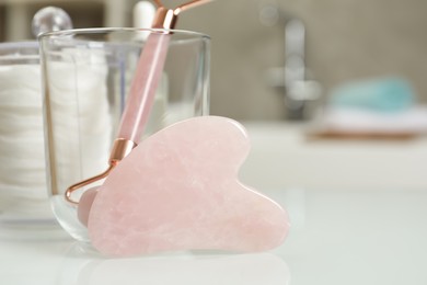 Photo of Rose quartz gua sha tool, natural face roller and toiletries on white table in bathroom, closeup. Space for text