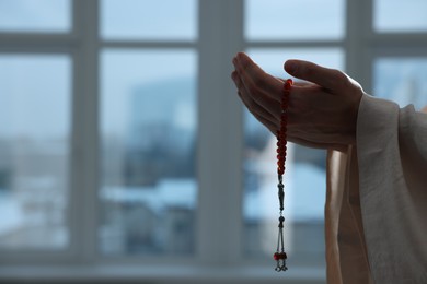 Muslim man with misbaha praying near window indoors, closeup. Space for text