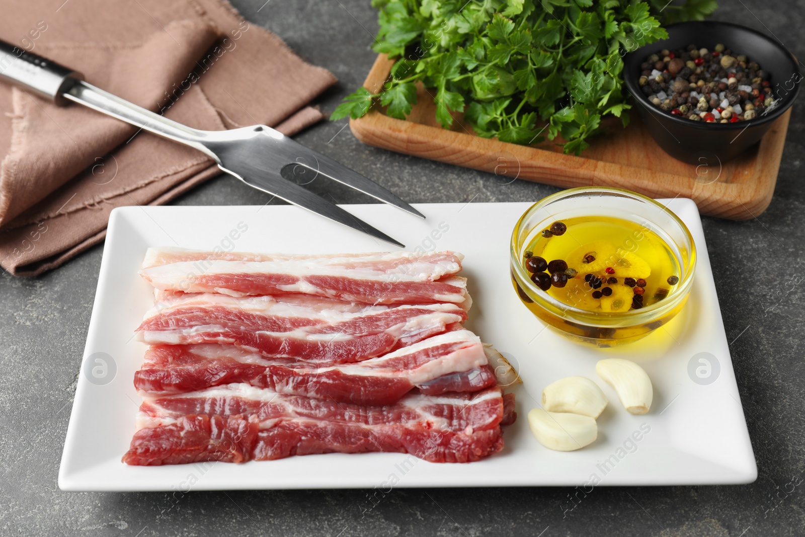 Photo of Pieces of raw pork belly, oil, parsley and garlic on grey textured table