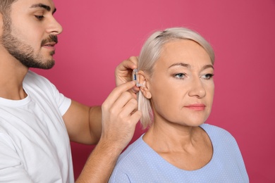 Young man putting hearing aid in mother's ear on color background