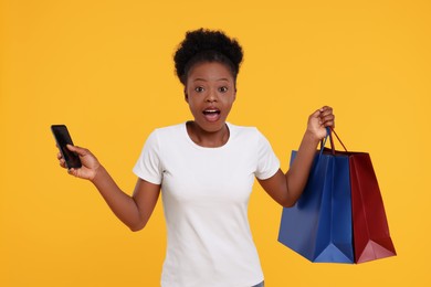 Photo of Shocked young woman with shopping bags and smartphone on orange background