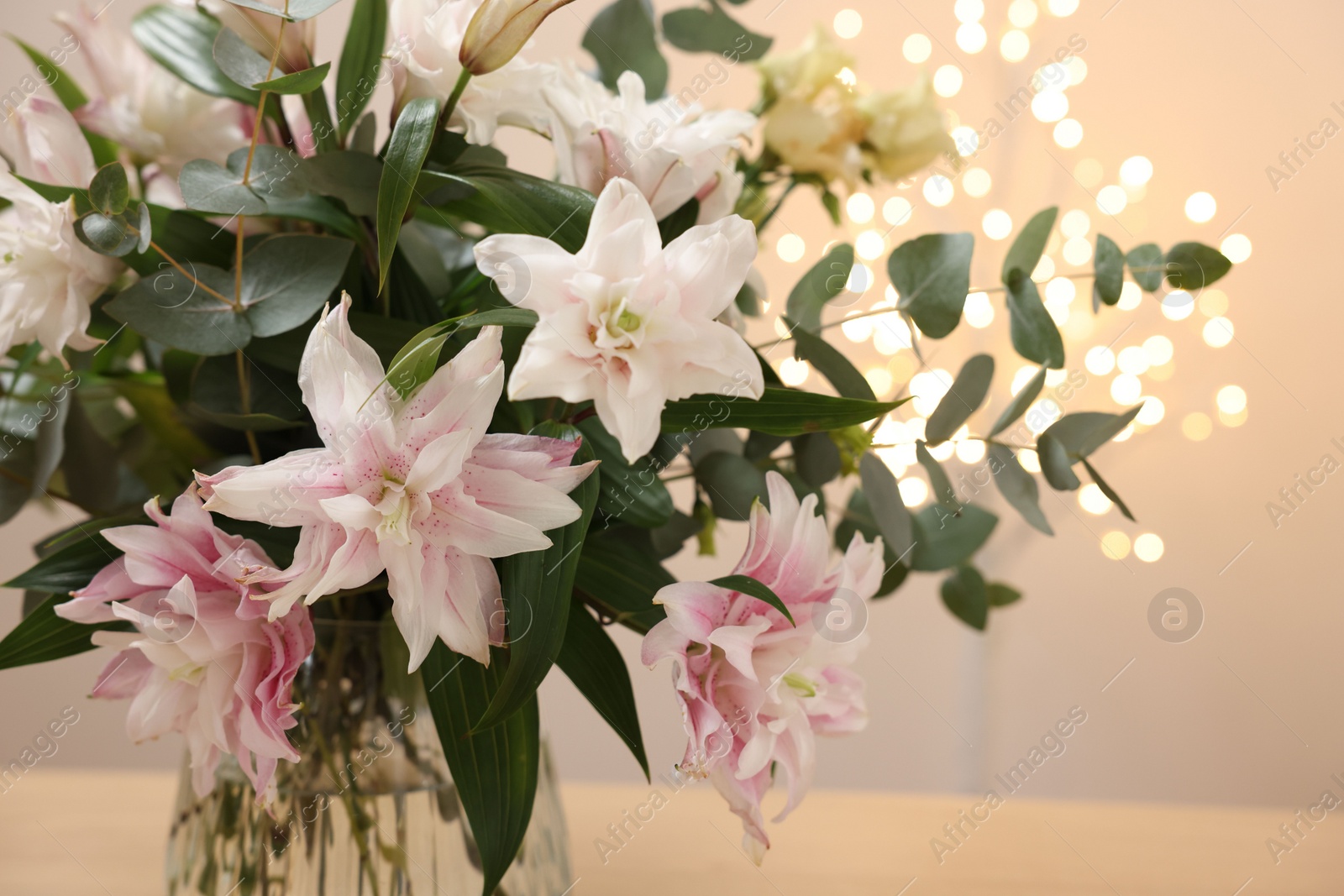 Photo of Bouquet of beautiful lily flowers in vase against beige background with blurred lights, closeup