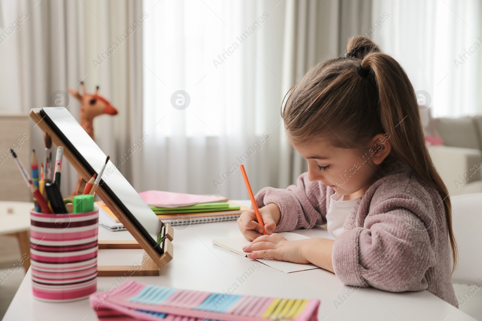 Photo of Adorable little girl doing homework with tablet at table indoors