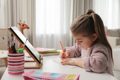 Adorable little girl doing homework with tablet at table indoors