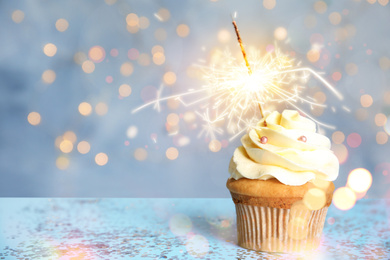 Image of Delicious birthday cupcake with sparkler on blue table against blurred lights. Space for text