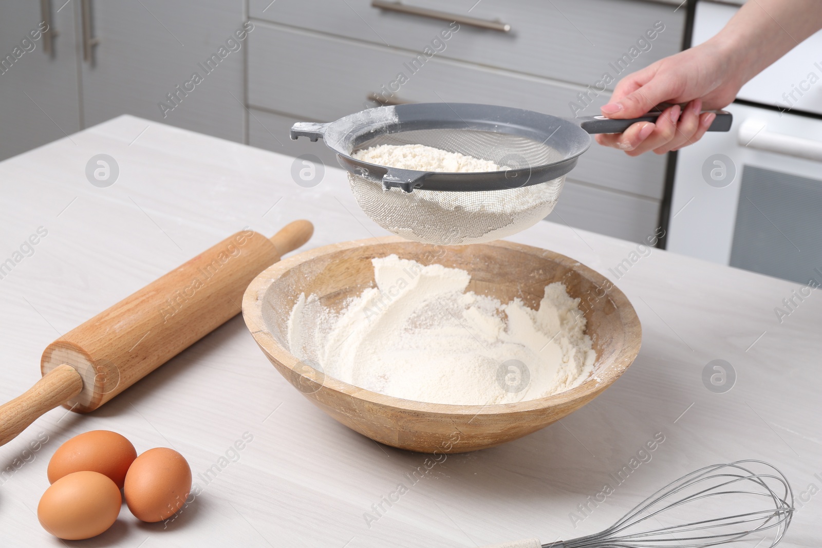 Photo of Woman sieving flour into bowl at white wooden table in kitchen, closeup