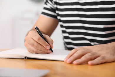 Young man writing in notebook at wooden table, closeup