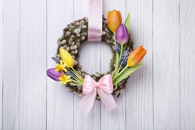 Willow wreath with different beautiful flowers and pink bow hanging on white wooden background