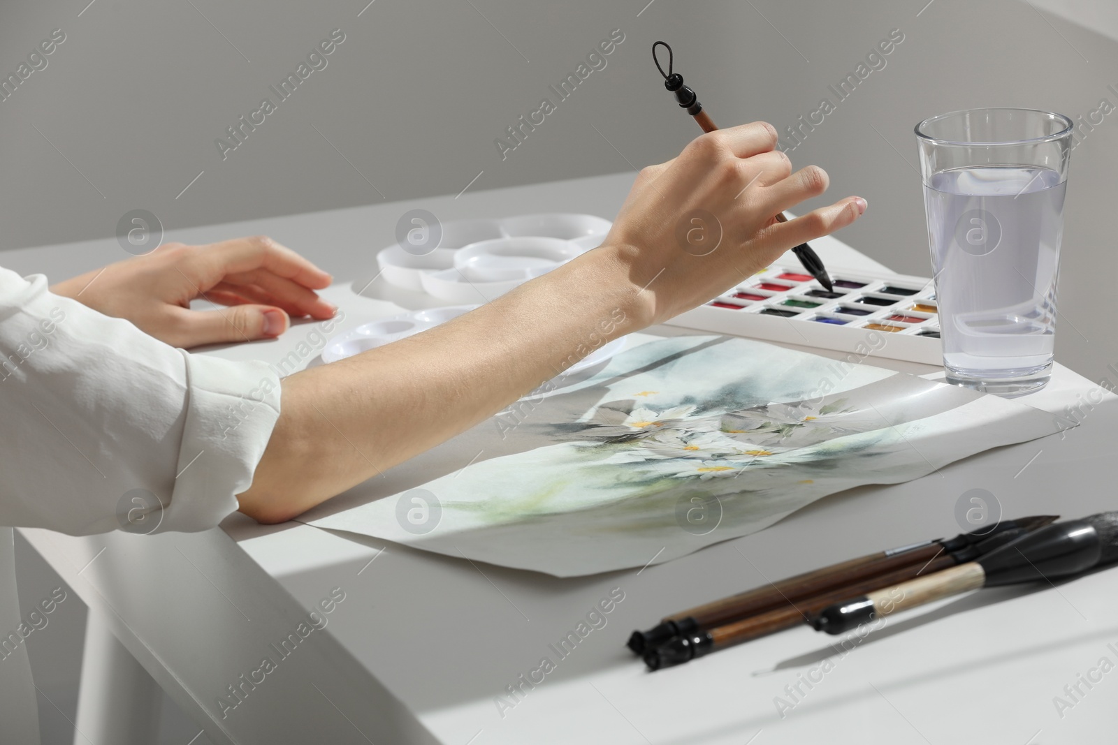 Photo of Woman painting flowers with watercolor at white table indoors, closeup. Creative artwork