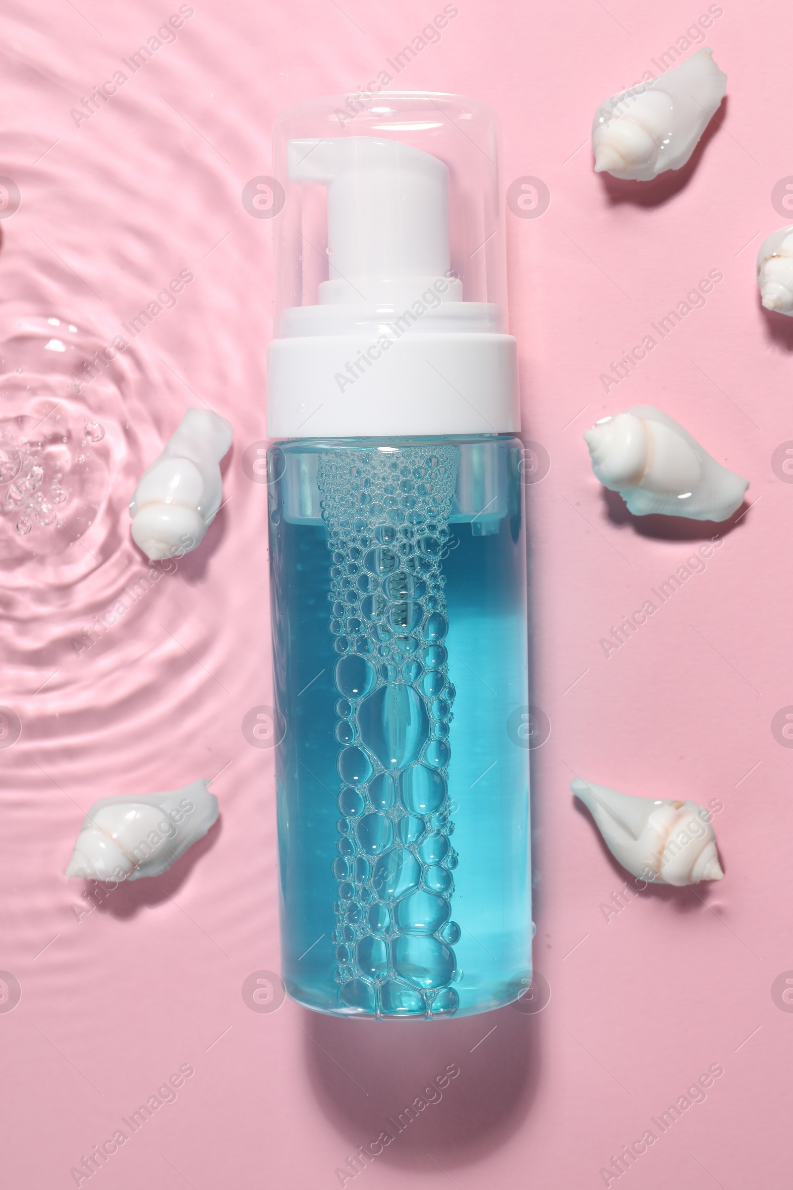 Photo of Bottle of facial cleanser and seashells in water against pink background, flat lay