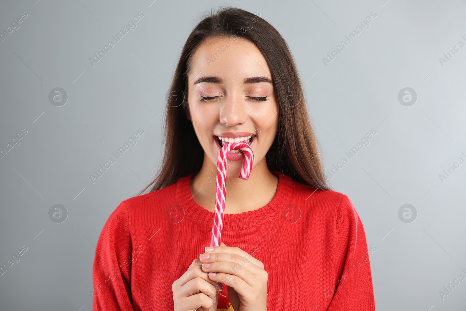 Photo of Young woman in Christmas sweater eating candy cane on grey background