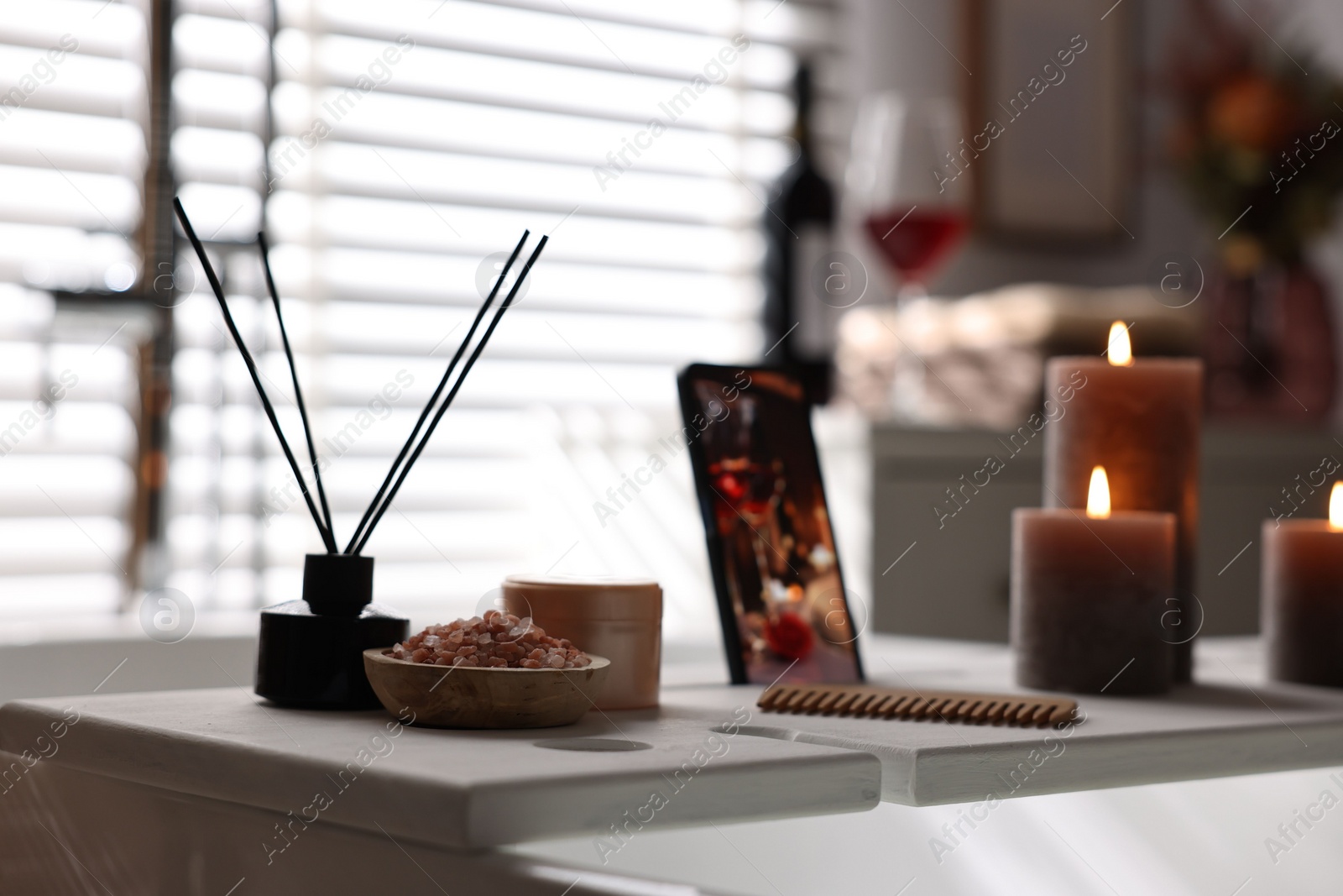 Photo of White wooden tray with aroma diffuser, beauty products and burning candles on bath tub in bathroom