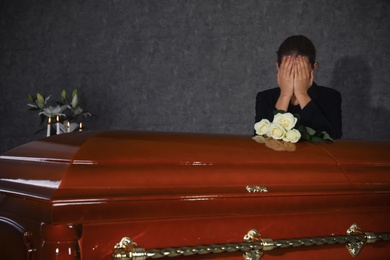 Photo of Sad young woman mourning near casket with white roses in funeral home
