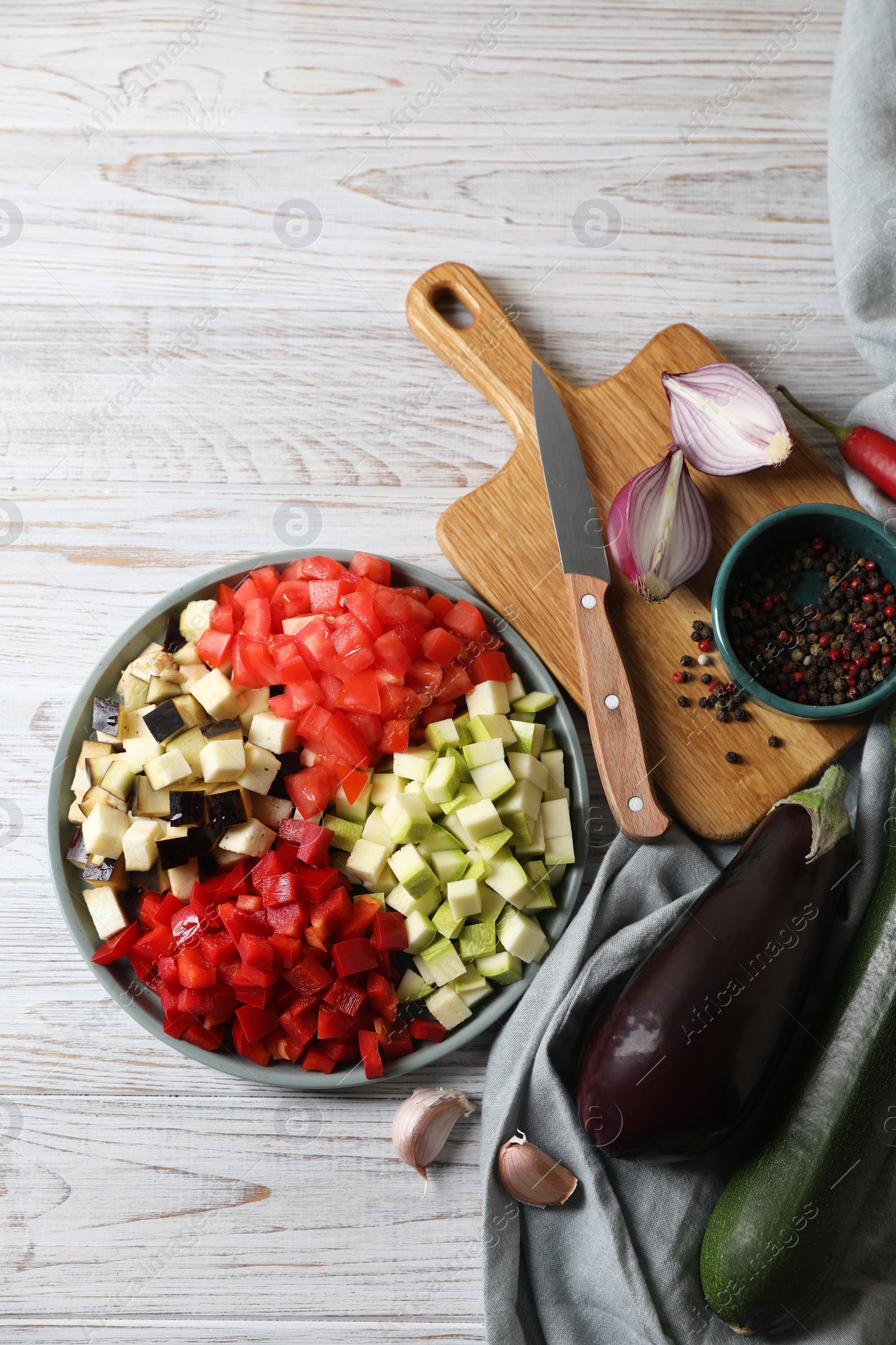 Photo of Cooking delicious ratatouille. Fresh ripe vegetables, plate and knife on white wooden table, flat lay