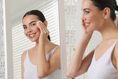 Beautiful woman removing makeup with cotton pad near mirror indoors