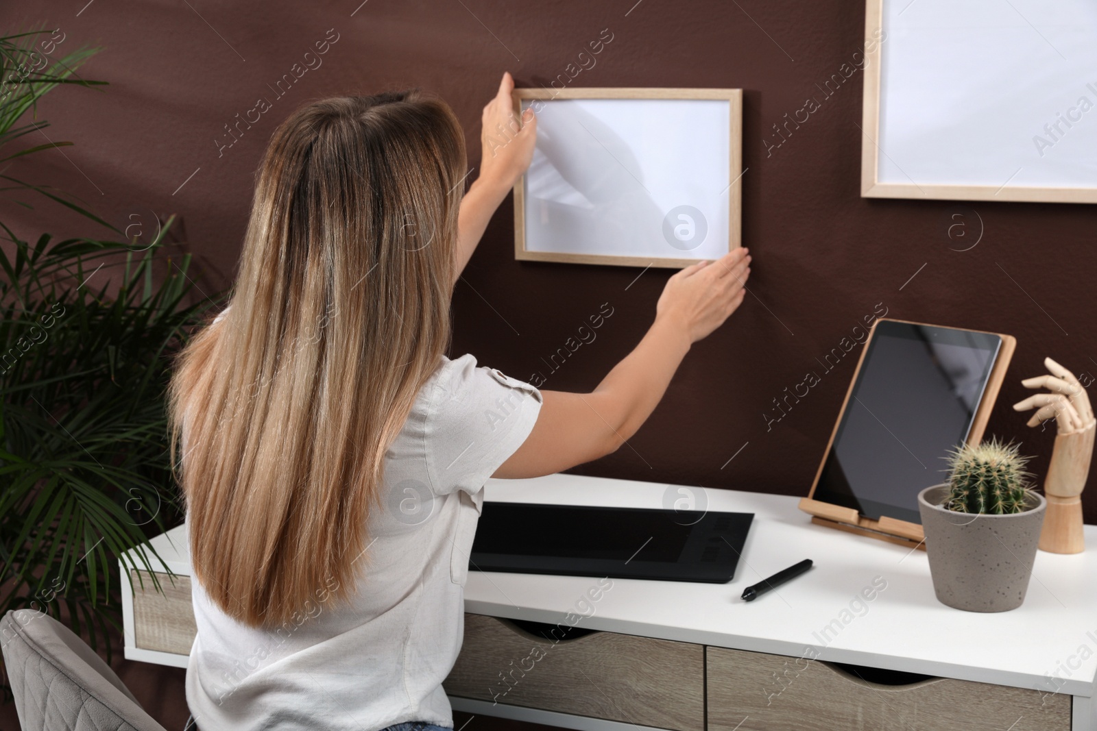 Photo of Young woman hanging empty frame on brown wall indoors. Mockup for design