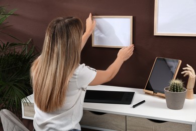 Young woman hanging empty frame on brown wall indoors. Mockup for design