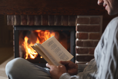 Photo of Man reading book near burning fireplace at home, closeup