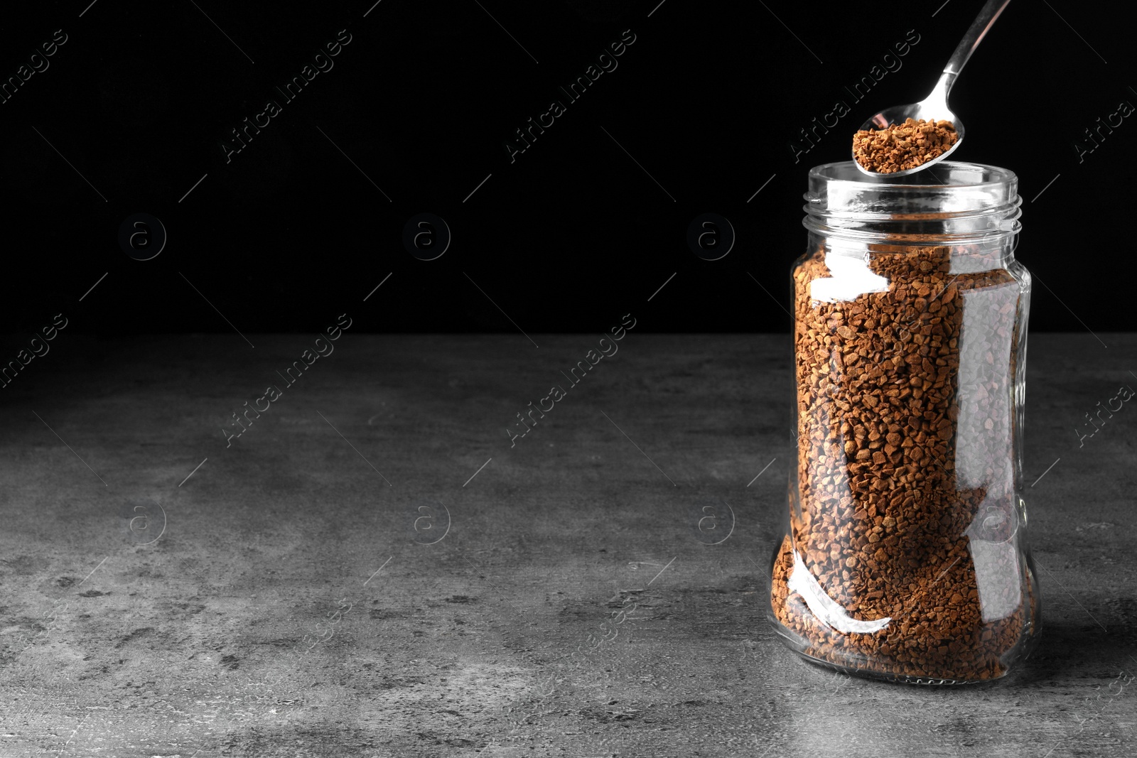 Photo of Instant coffee and spoon above glass jar on grey table against black background. Space for text