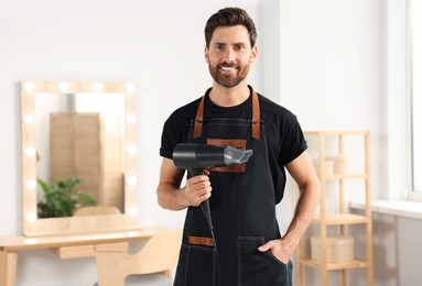 Smiling hairdresser in apron holding dryer near vanity mirror in salon