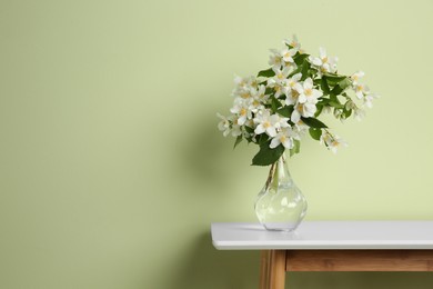 Photo of Bouquet of beautiful jasmine flowers in glass vase on table near green wall, space for text