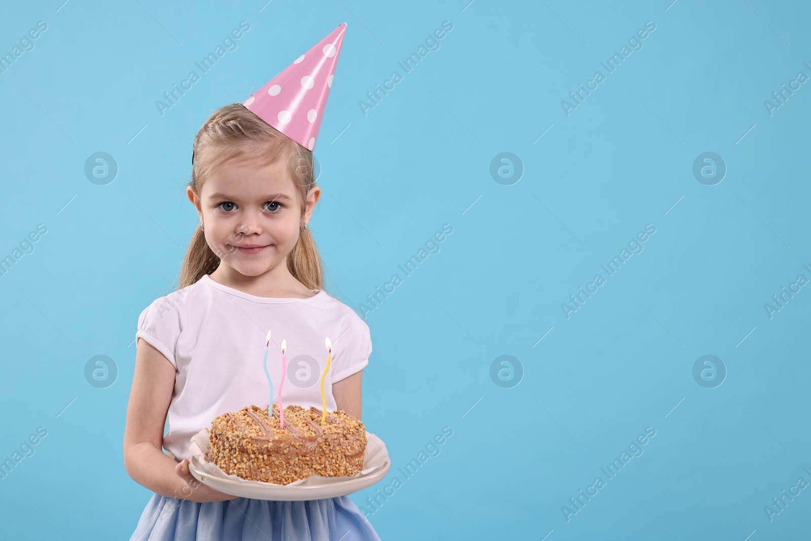 Photo of Birthday celebration. Cute little girl in party hat holding tasty cake with burning candles on light blue background, space for text