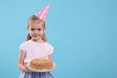 Birthday celebration. Cute little girl in party hat holding tasty cake with burning candles on light blue background, space for text