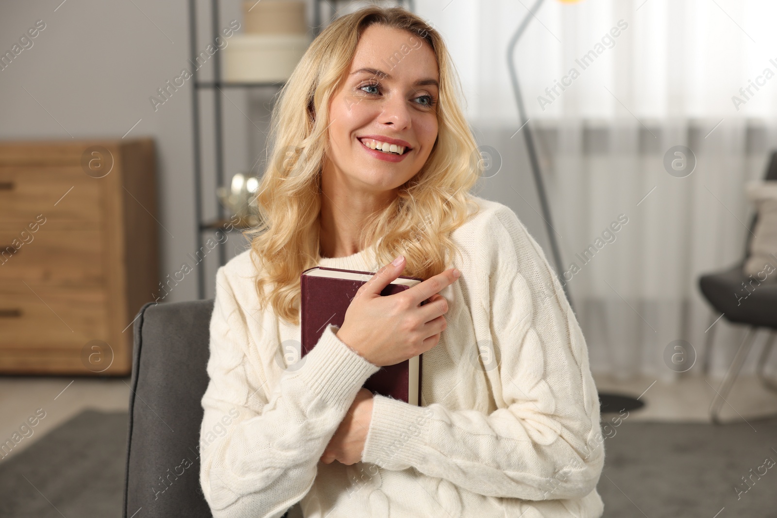 Photo of Happy woman in stylish warm sweater holding book at home