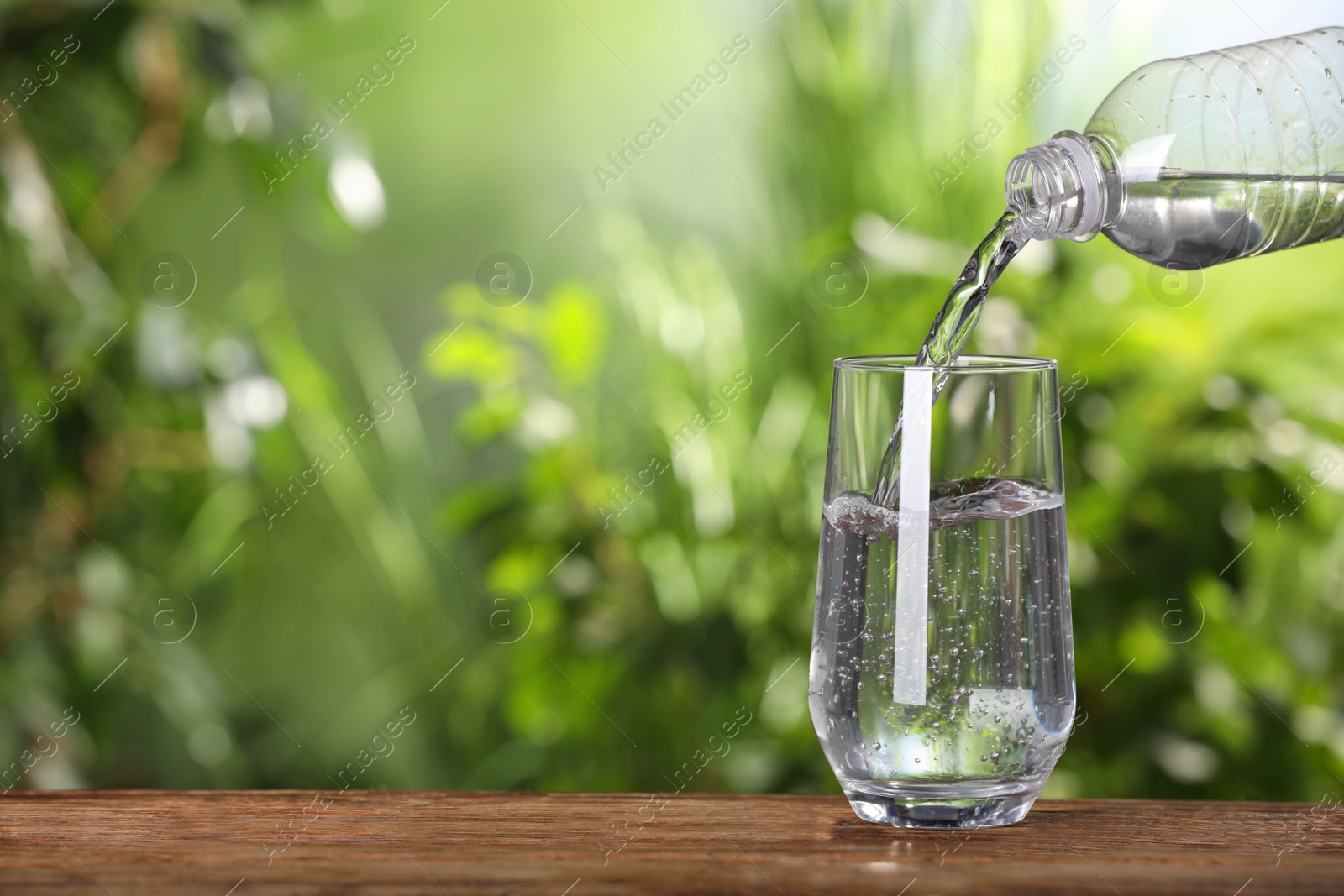 Photo of Pouring water from bottle into glass on wooden table outdoors, closeup. Space for text