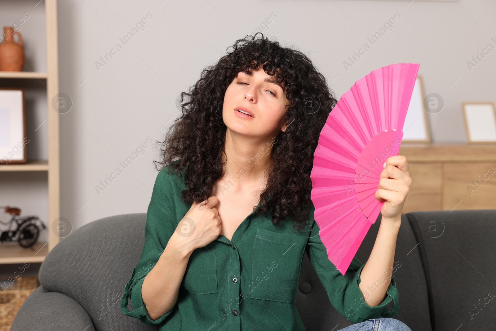 Photo of Young woman waving pink hand fan to cool herself on sofa at home